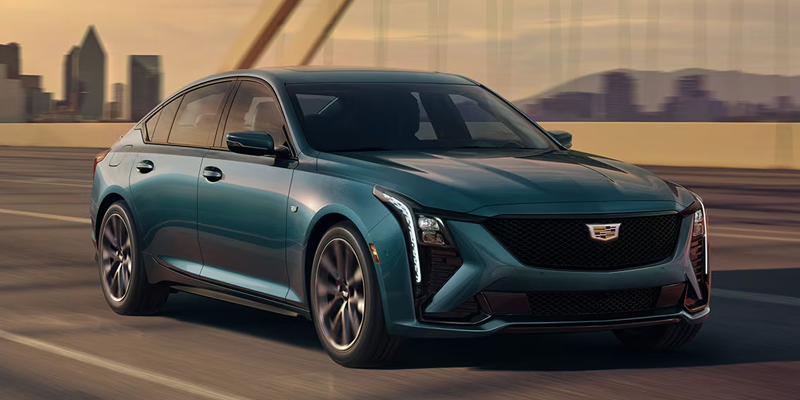 Blue-green Cadillac sedan driving on a bridge with a city skyline in the background