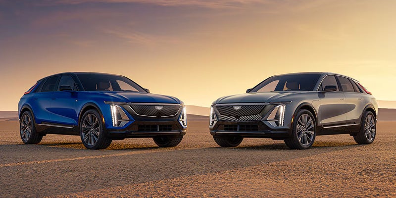 Blue and silver Cadillac SUVs parked side by side in a desert landscape at sunset