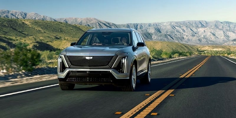 Silver Cadillac SUV driving on an open highway with mountains in the background.